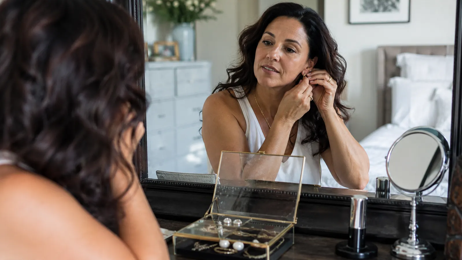 Woman getting ready in the morning, choosing earrings from an open glass-lid jewelry box on her dresser