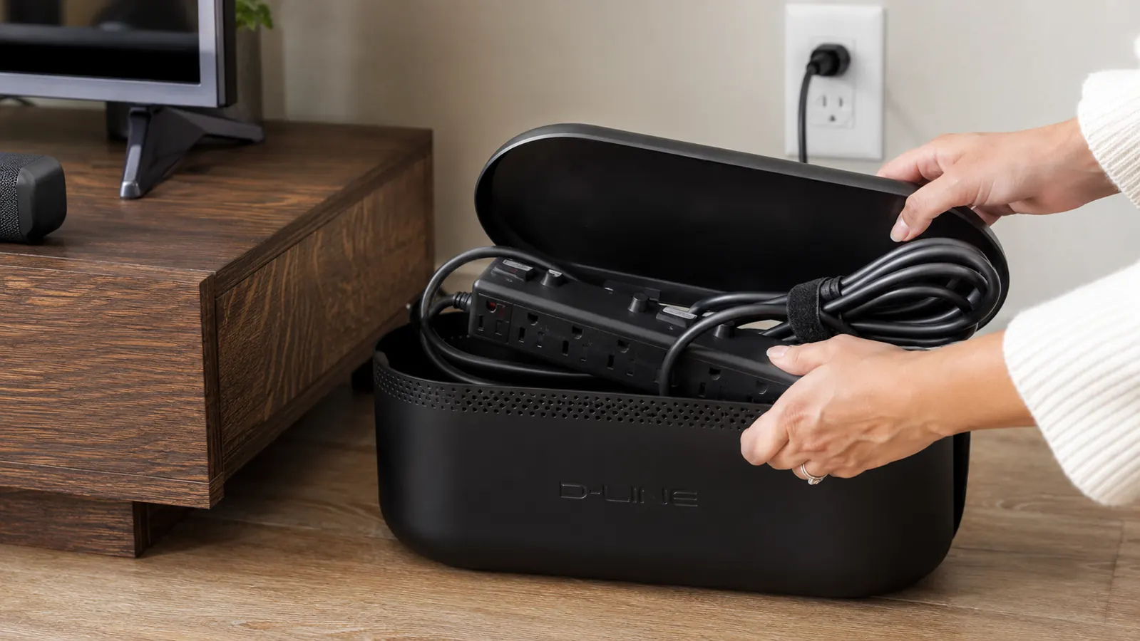 Hand tucking a power strip and cords into an open D-Line cable management box behind a TV stand