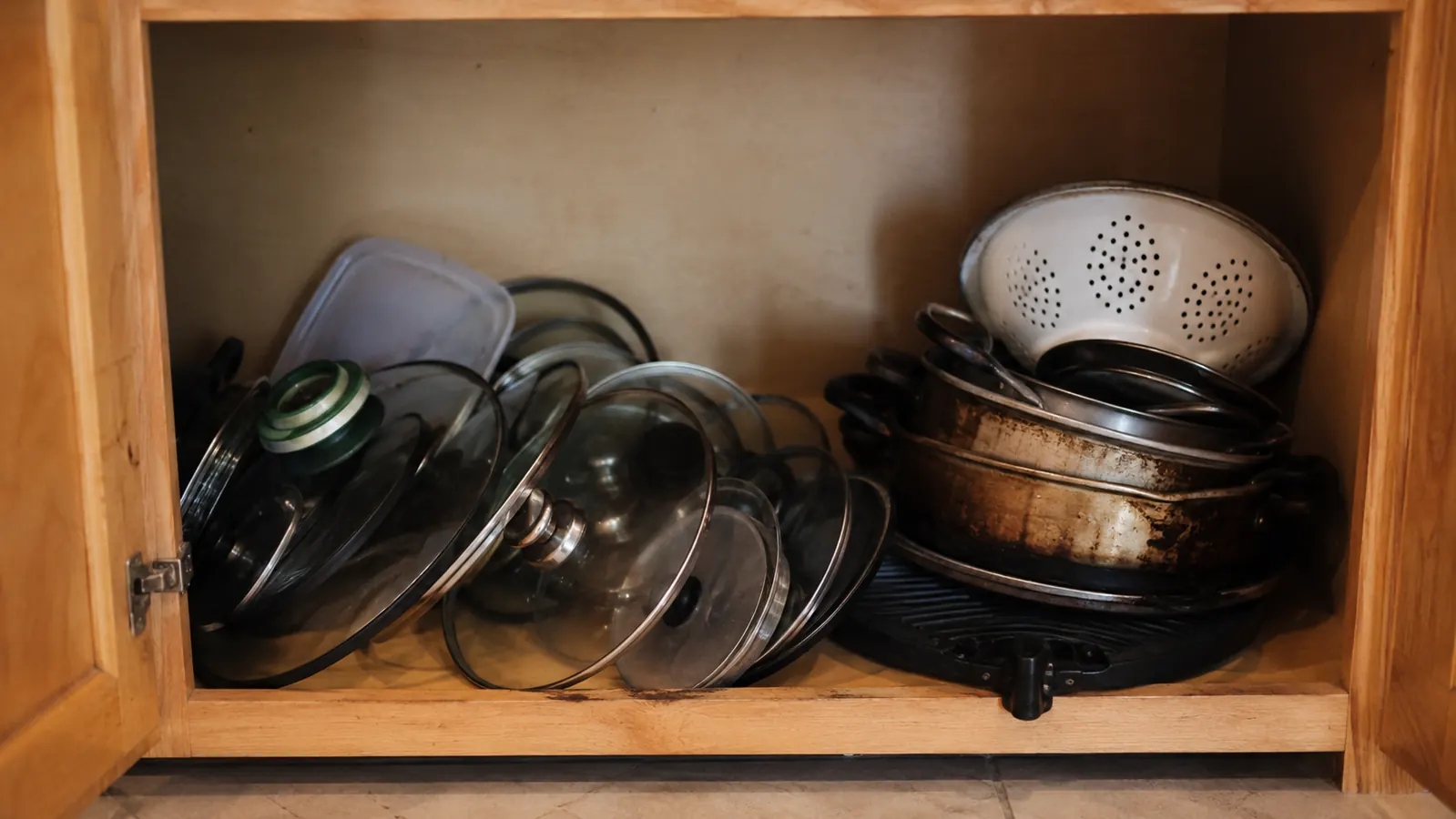 Cluttered lower cabinet with mismatched pot lids stacked loosely and sliding against each other